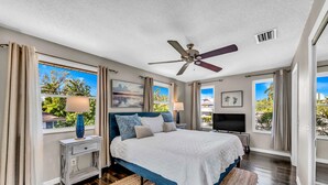 A bedroom with a bed, ceiling fan, TV, and large windows with curtains. - You, Me, and the Sea AMI - Elevated Beach Home with Pool and Spa Steps from Beac (Holmes Beach)