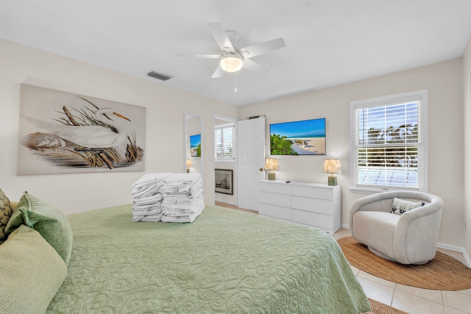 A bedroom with a bed, a chair, a vanity with a mirror, a ceiling fan, and artwork on the walls.