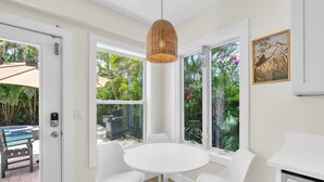 A room with a round white table and four white chairs, large windows with a view of a pool and tropical plants, and a hanging woven light fixture.
