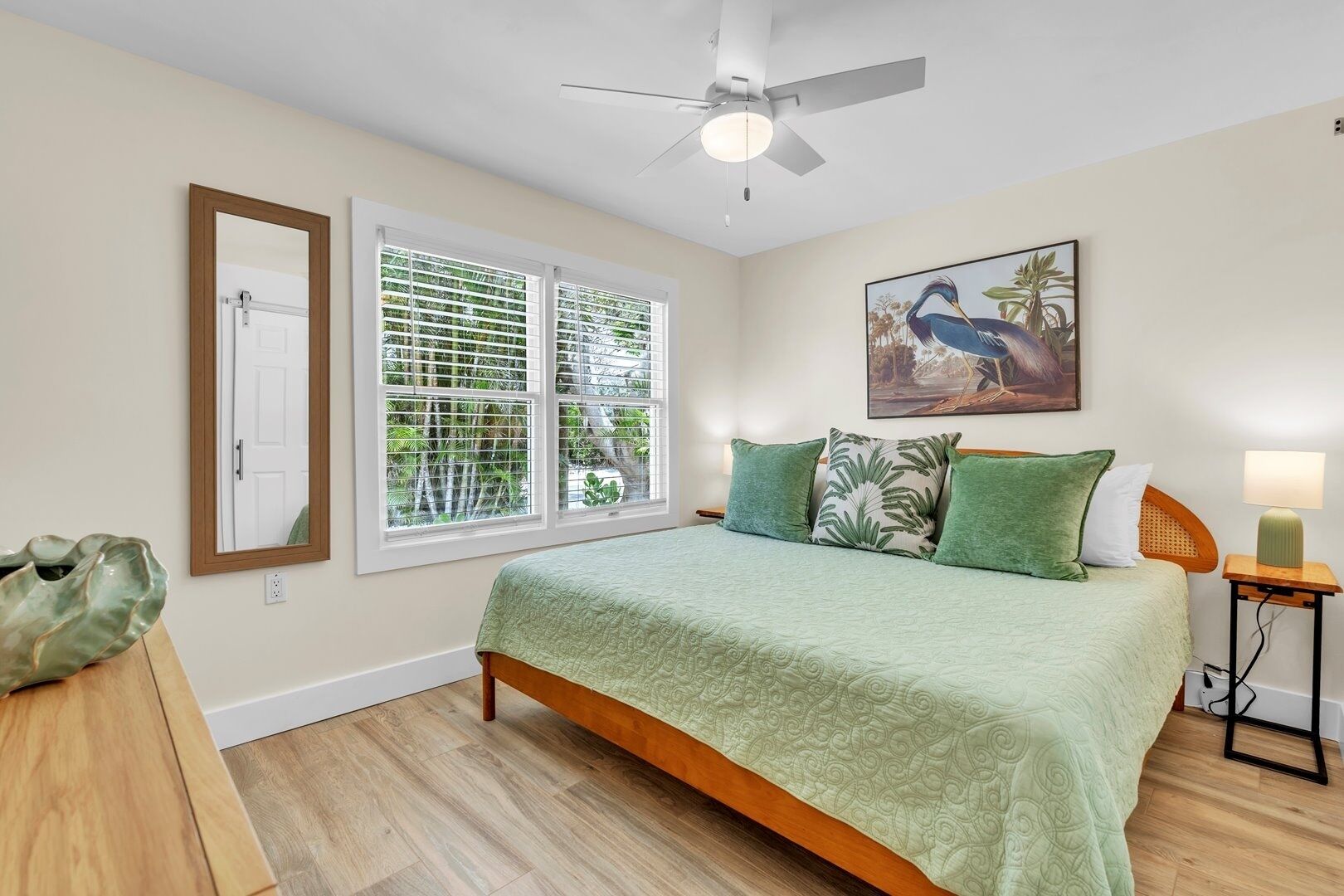 A bedroom with a bed, a ceiling fan, a window with blinds, a door, a wall-mounted painting, and a bedside table with a lamp.