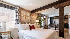 Een ruime slaapkamer met een groot bed, een nachtkastje, een lamp en uitzicht op de keuken.
