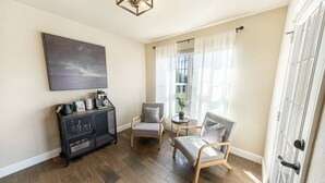 Ein Zimmer mit Holzboden, einem schwarzen Metallschrank mit Gegenständen darauf, zwei grauen Stühlen, einem kleinen runden Tisch mit einer Pflanze und einem großen Fenster mit weißen Vorhängen.
