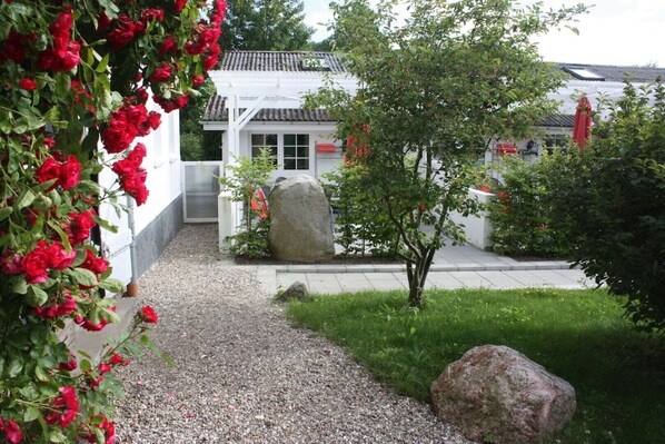 Ein Garten mit einem Kiesweg, der zu einem weißen Gebäude mit einem Ziegeldach führt. Rote Blumen, ein großer Felsen und ein kleiner Baum befinden sich im Garten.