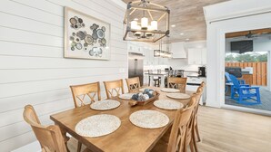 A dining area with a wooden table set for six, surrounded by wooden chairs. A chandelier hangs above, and a framed artwork is on the wall.