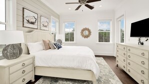A bedroom with a bed, dresser, ceiling fan, artwork, and large windows.