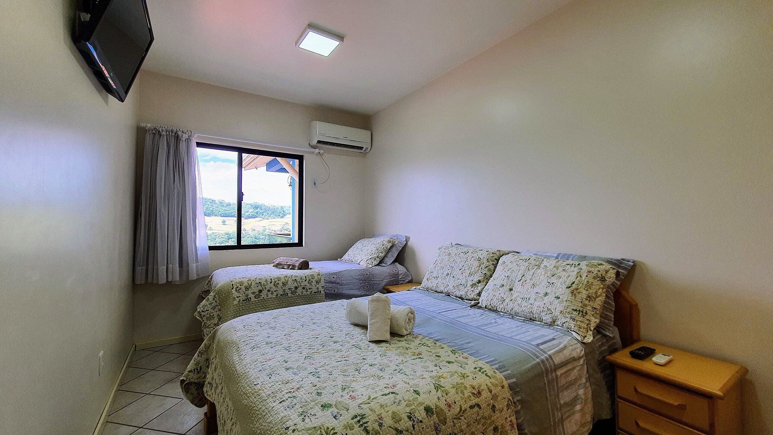 A bedroom with two beds, a TV, an air conditioner, and a window with curtains.