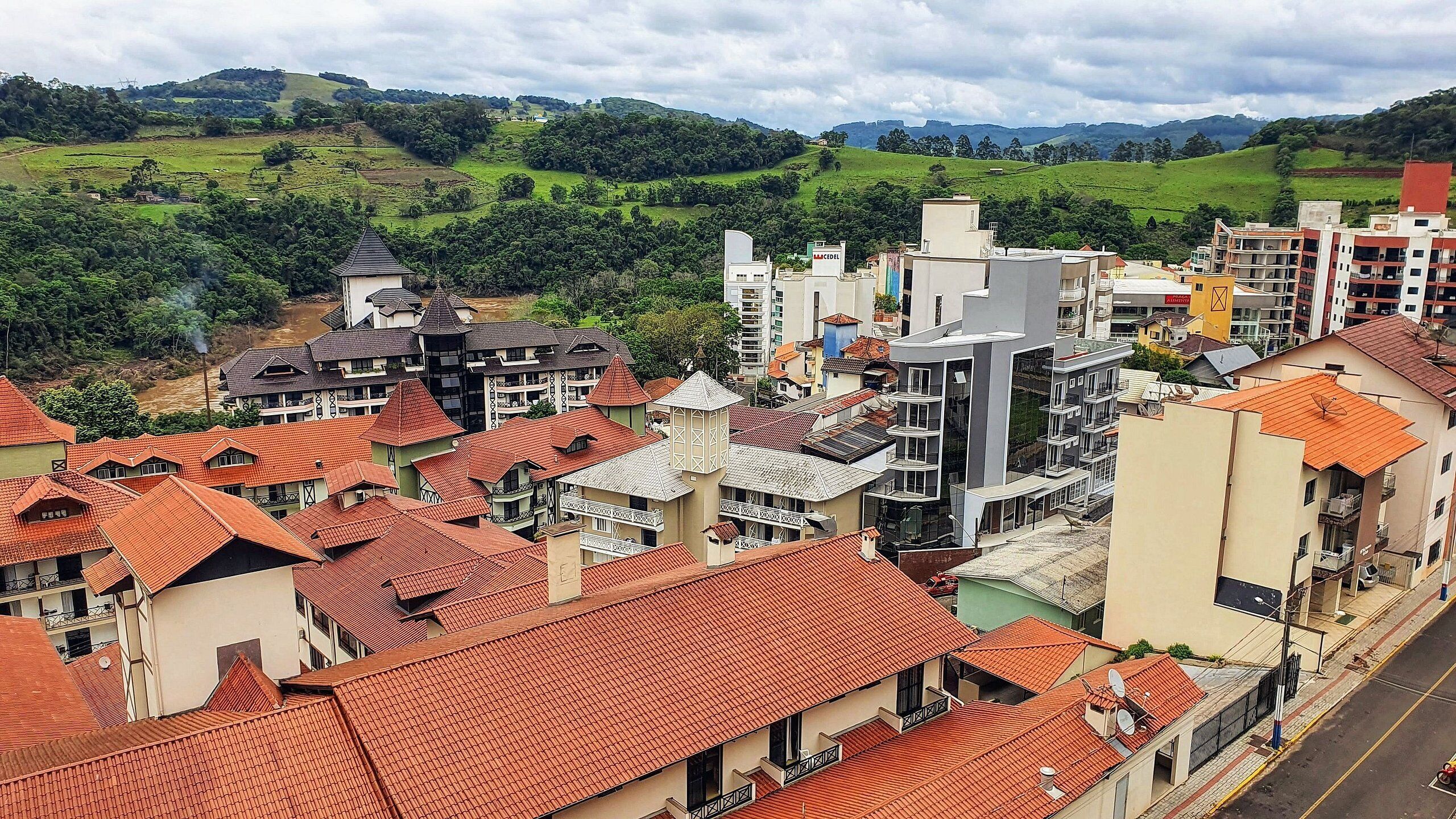 A residential area with red-tiled roofs, modern buildings, and a hilly landscape in the background.