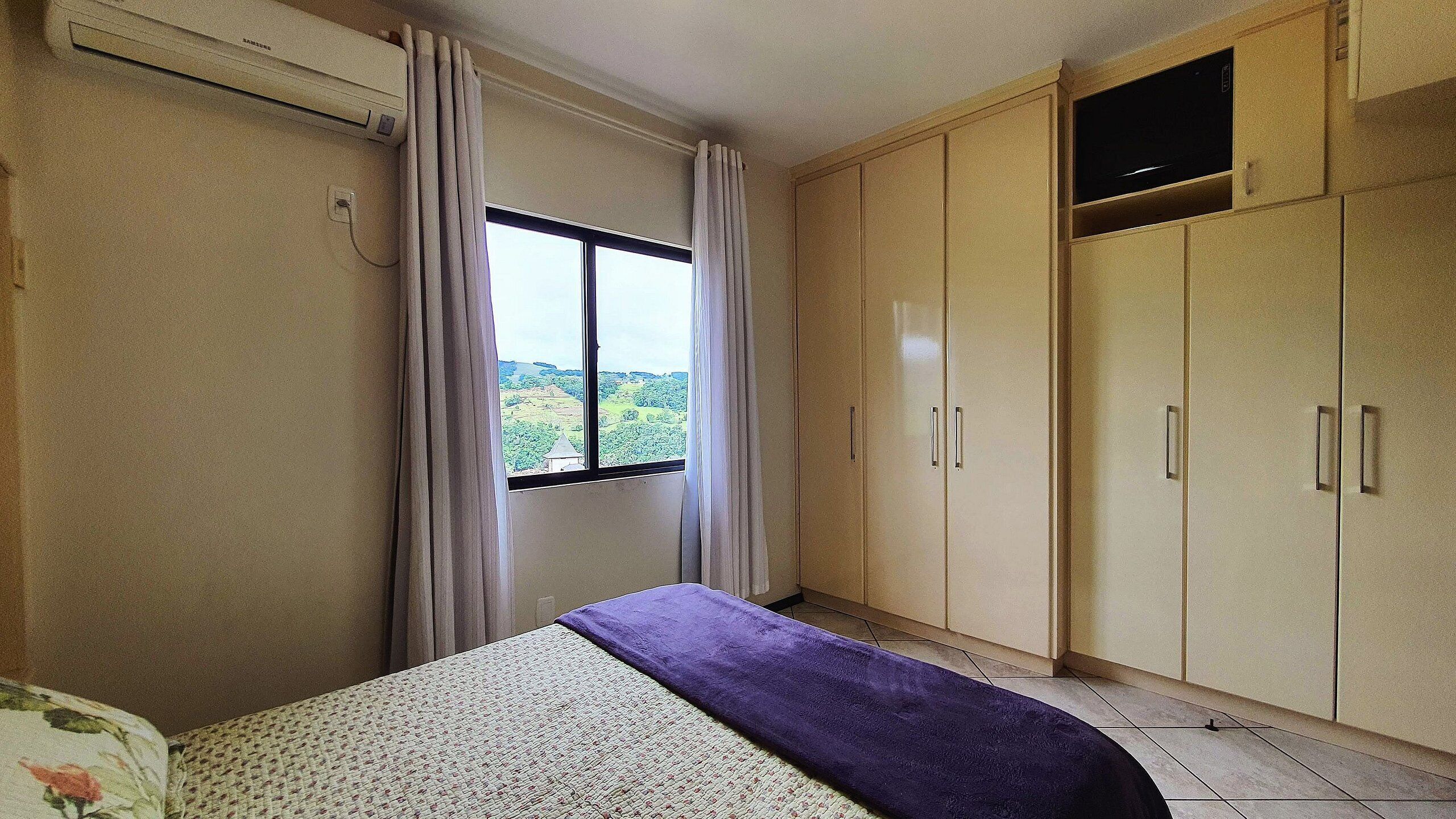 Une chambre à coucher avec un lit, une fenêtre avec des rideaux, une armoire et un téléviseur posé sur une étagère.