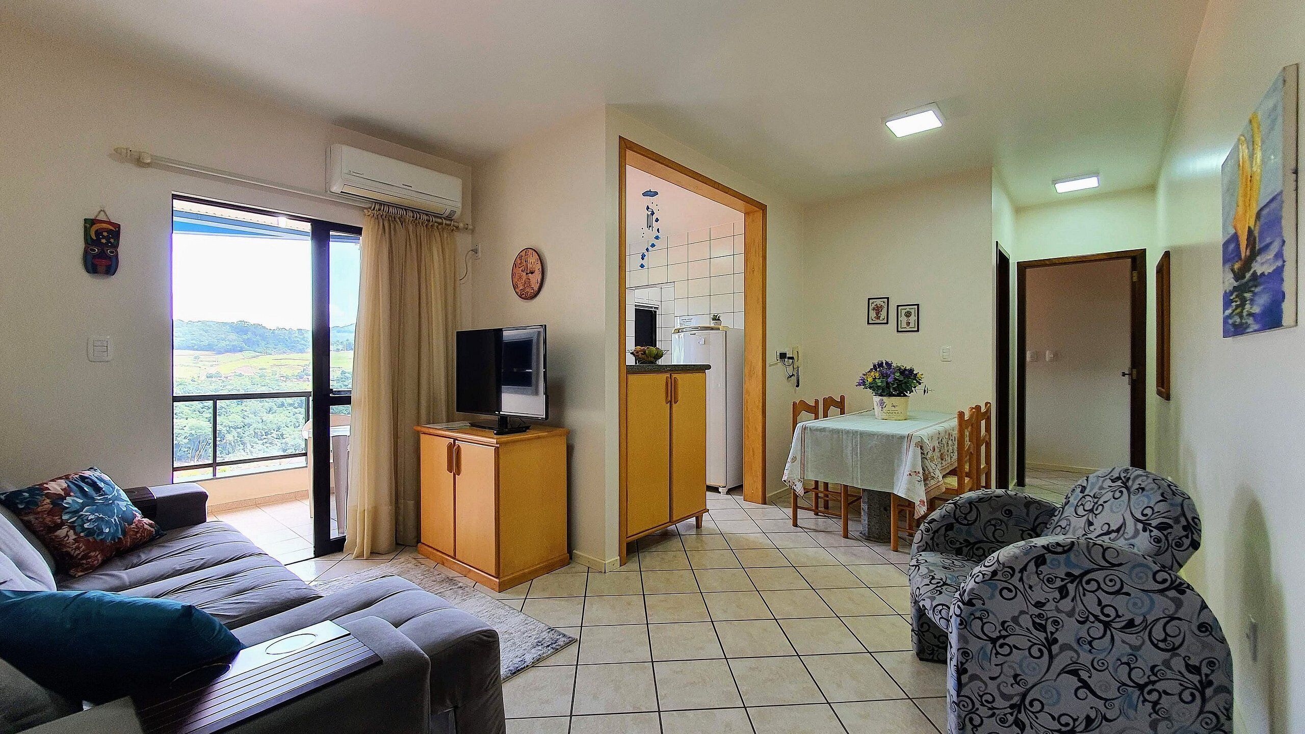 A living room with a sofa, a TV on a wooden cabinet, a dining area with a table and chairs, and a kitchen in the background.