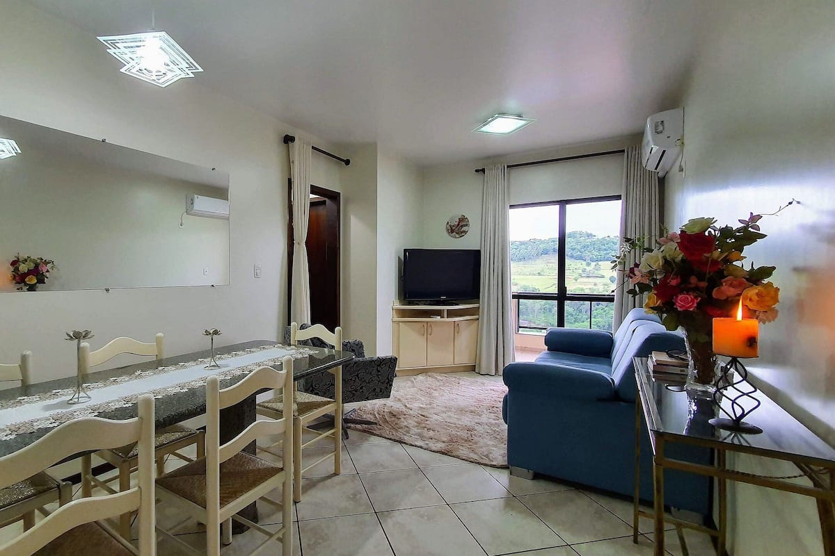 A dining area with a table and chairs, a TV, a sofa, and a window with a view.