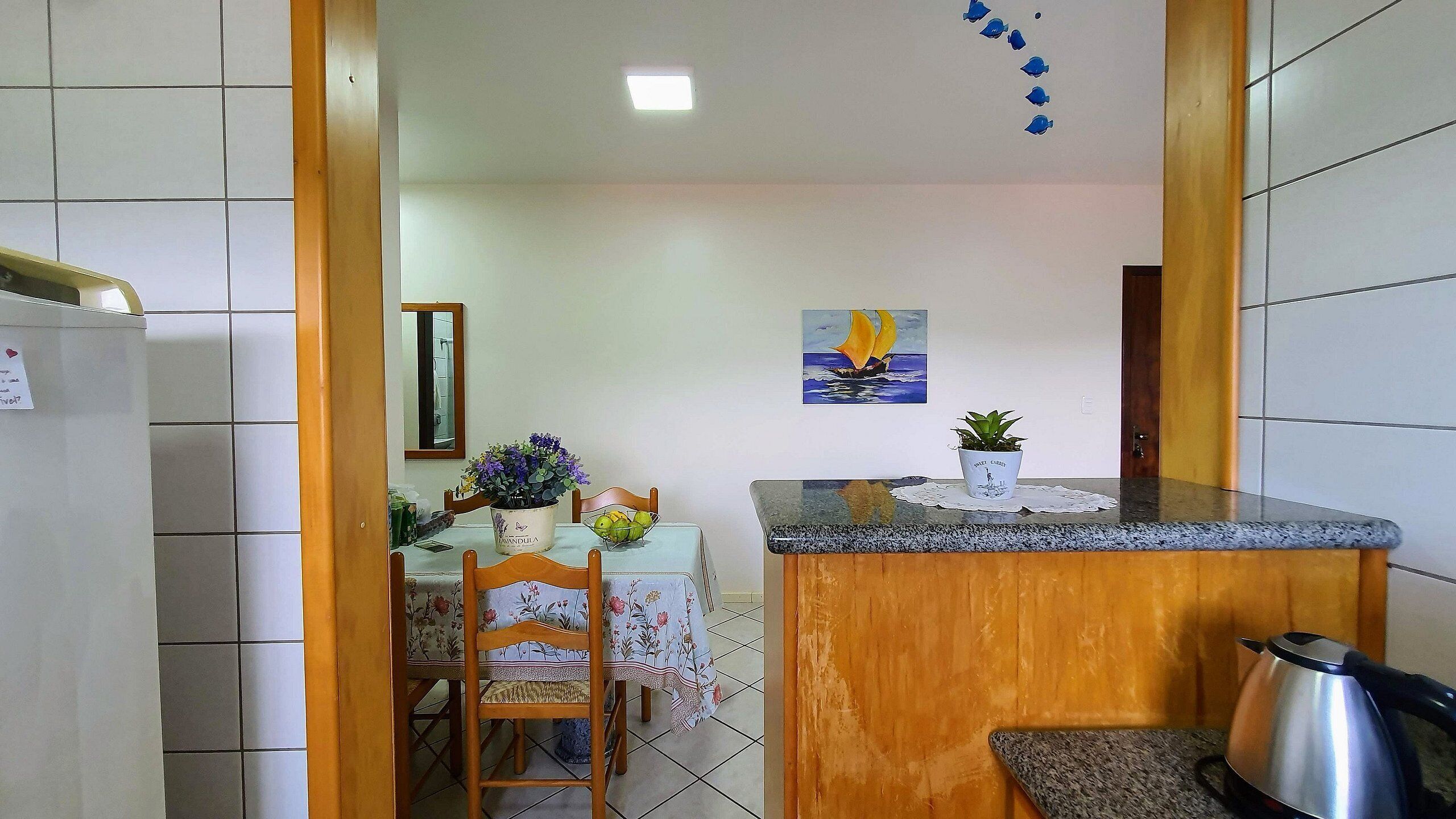 A kitchen with a tiled backsplash, a wooden countertop, a sink, and a stove. There is a dining table with chairs and a potted plant. A painting of a sailboat is on the wall.