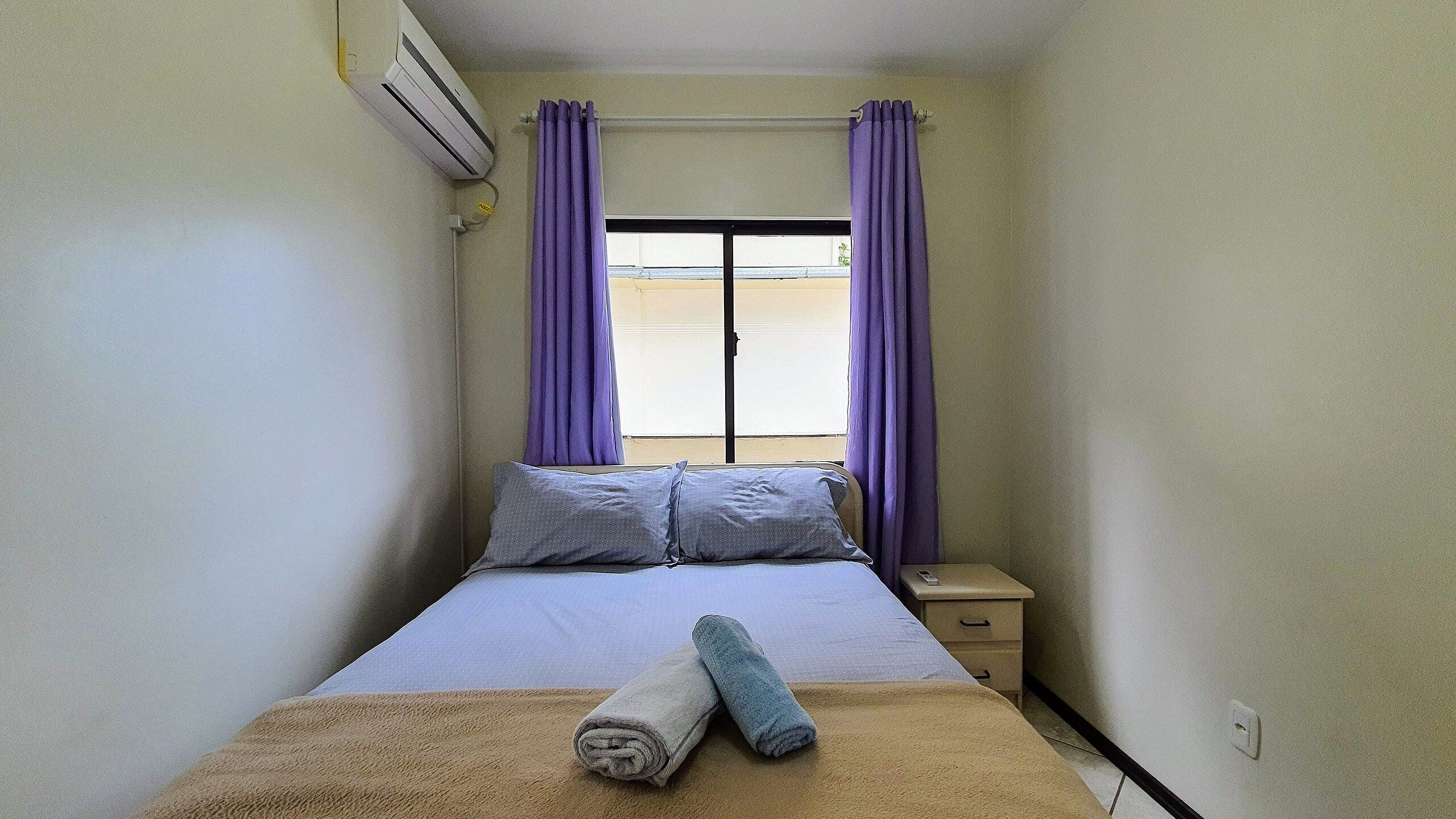 A small, simple room with a bed, a nightstand, and a window with purple curtains.