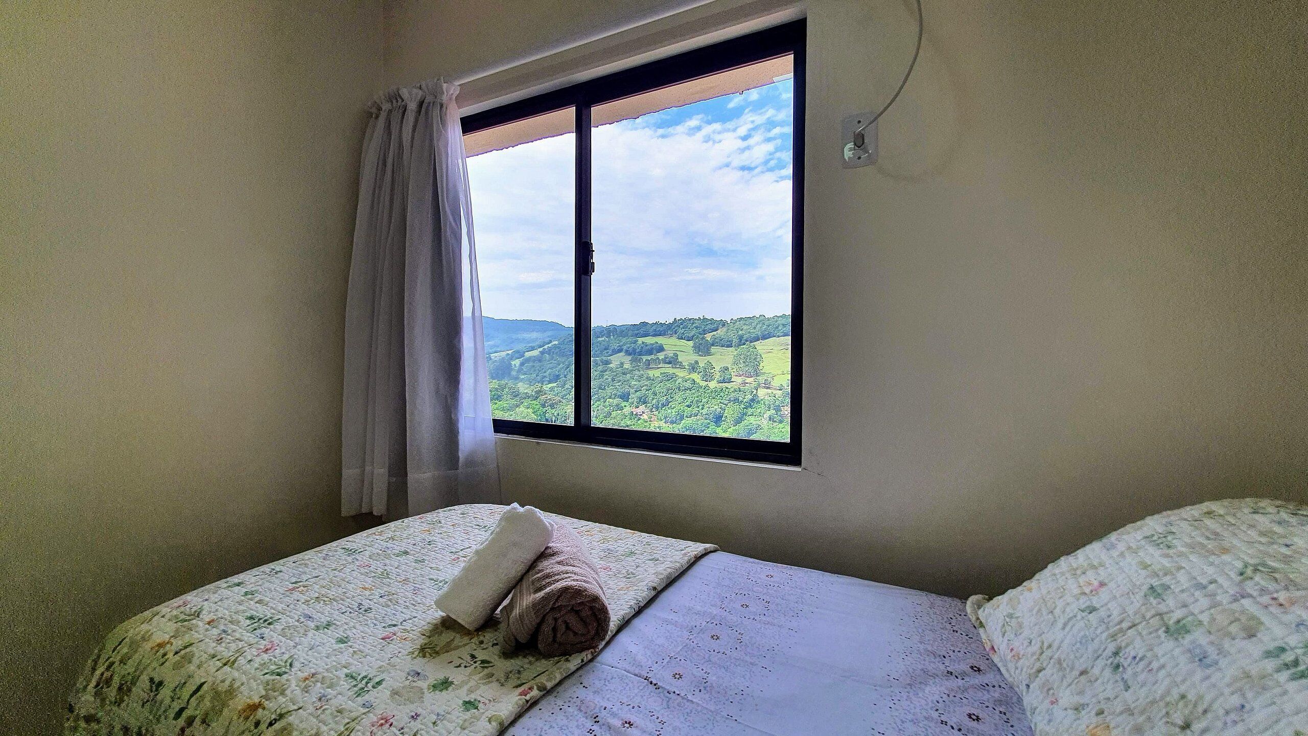 Un dormitorio con una cama, una ventana que ofrece vistas a un paisaje verde y un enchufe en la pared.