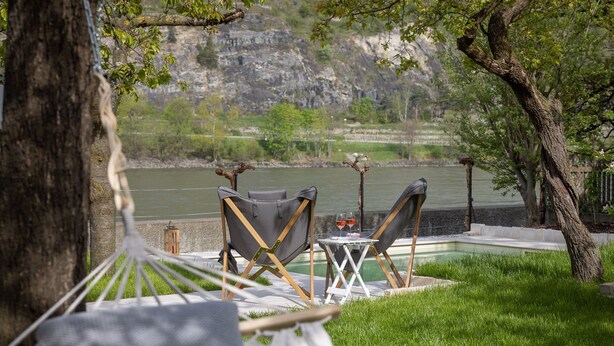 Una hamaca, dos sillas plegables, una mesita con vasos y vistas a un río y montañas.