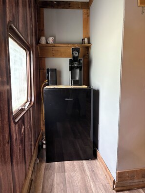 A compact kitchen with a black refrigerator, wooden shelves, a coffee machine, and a window with a view.