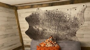 Ein Bett mit einer grau gemusterten Tagesdecke, ein dekoratives Kissen mit floralem Muster und ein schwanenförmiges Kissen.