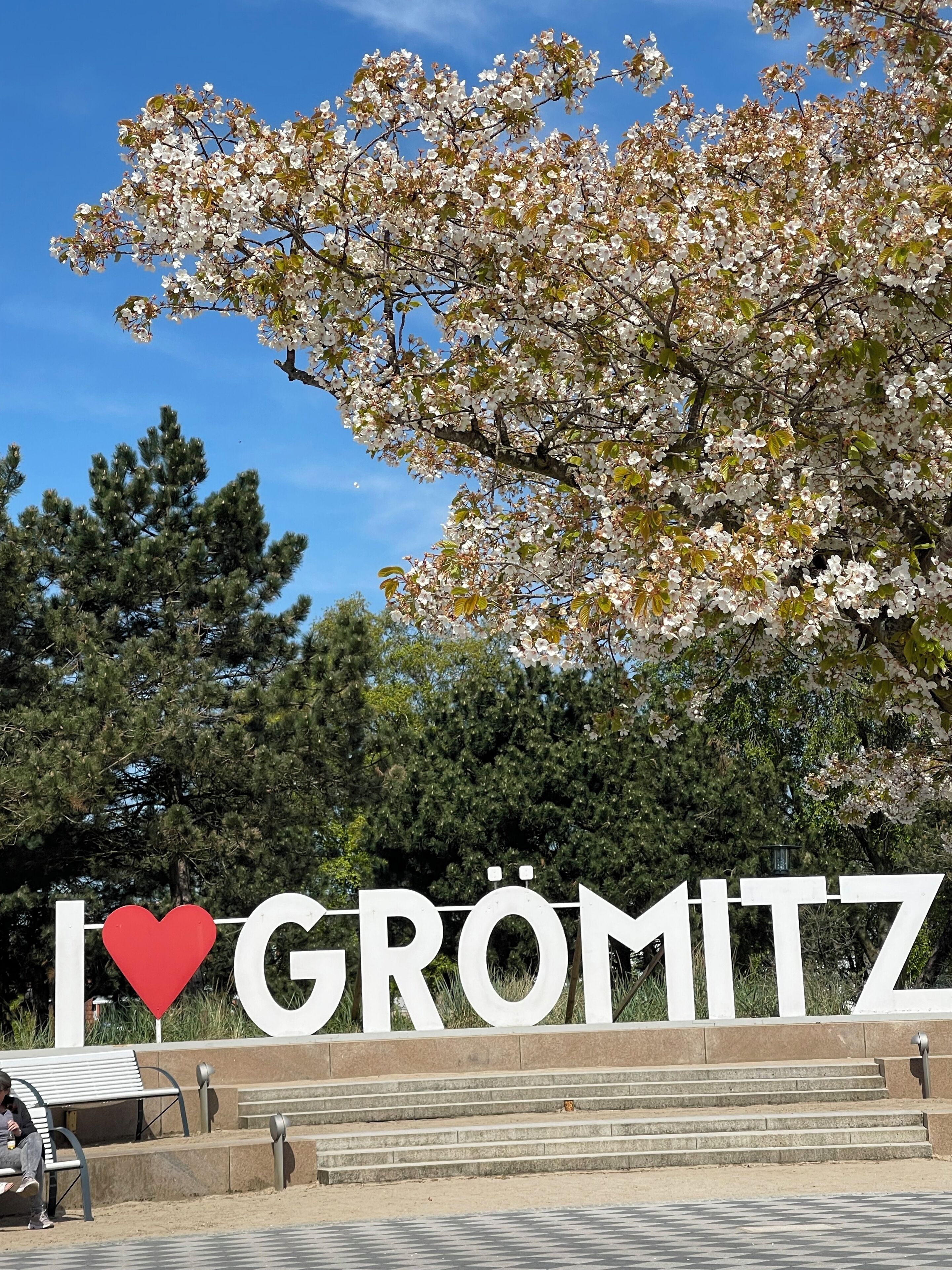 Ein Schild mit dem Aufdruck 'I ♥ GRÖMITZ' und einem Herzsymbol, im Vordergrund eine Bank und Treppen.