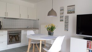 Una cocina moderna con armarios blancos, horno empotrado, una mesa de comedor con sillas, una planta en maceta y un televisor de pantalla plana sobre un mueble blanco.