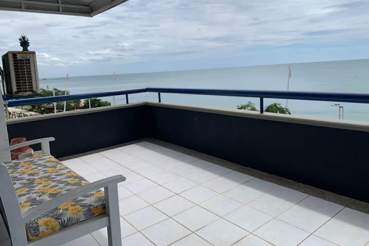 A balcony with a white chair and a table, overlooking a body of water and a distant cityscape.