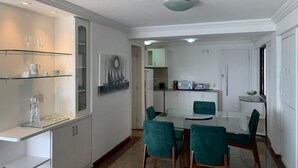 Un área de comedor moderna con una mesa de vidrio y sillas color turquesa, armarios blancos y una cocina al fondo.