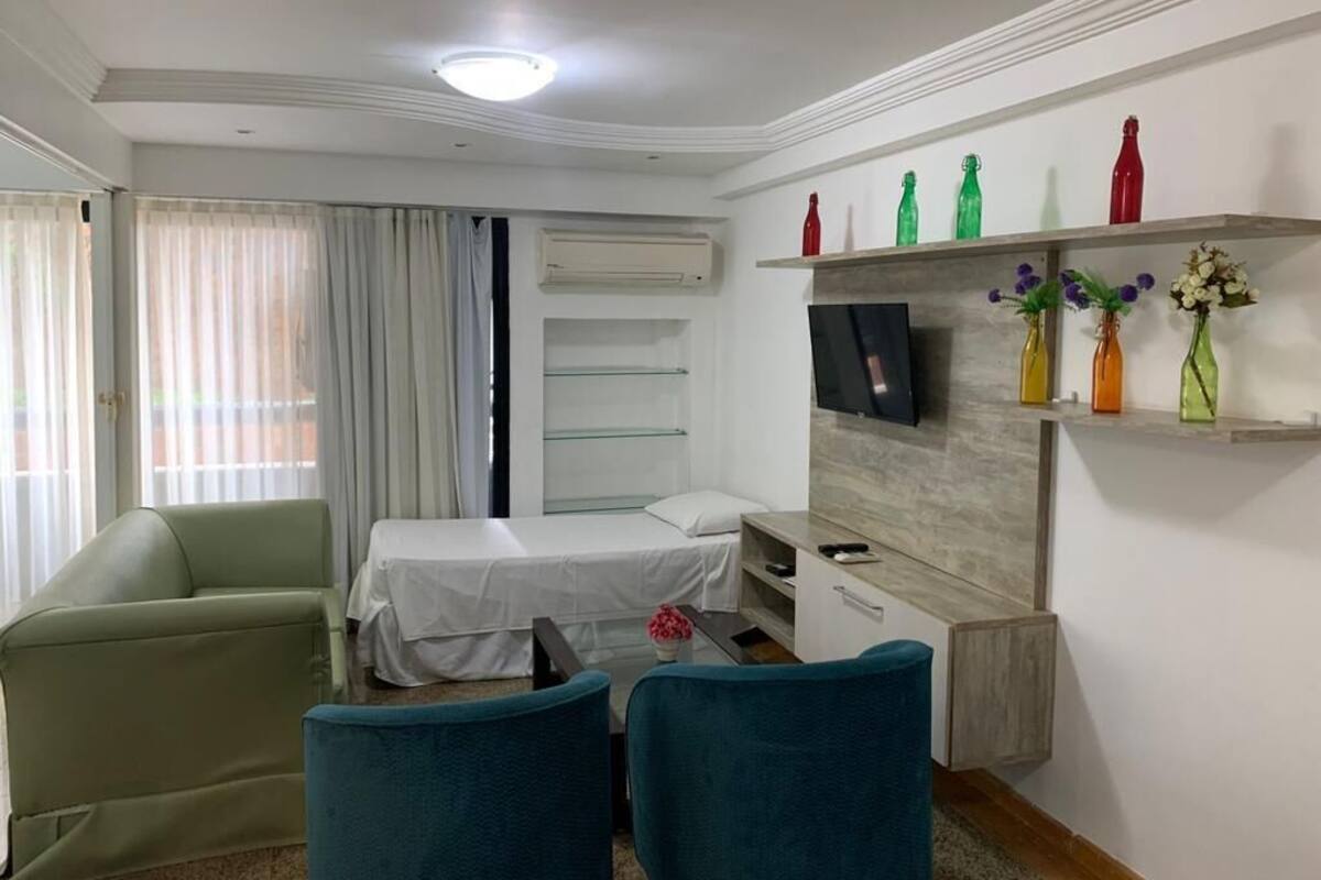A hotel room with a bed, a desk, a chair, a TV, and a shelf with decorative bottles.