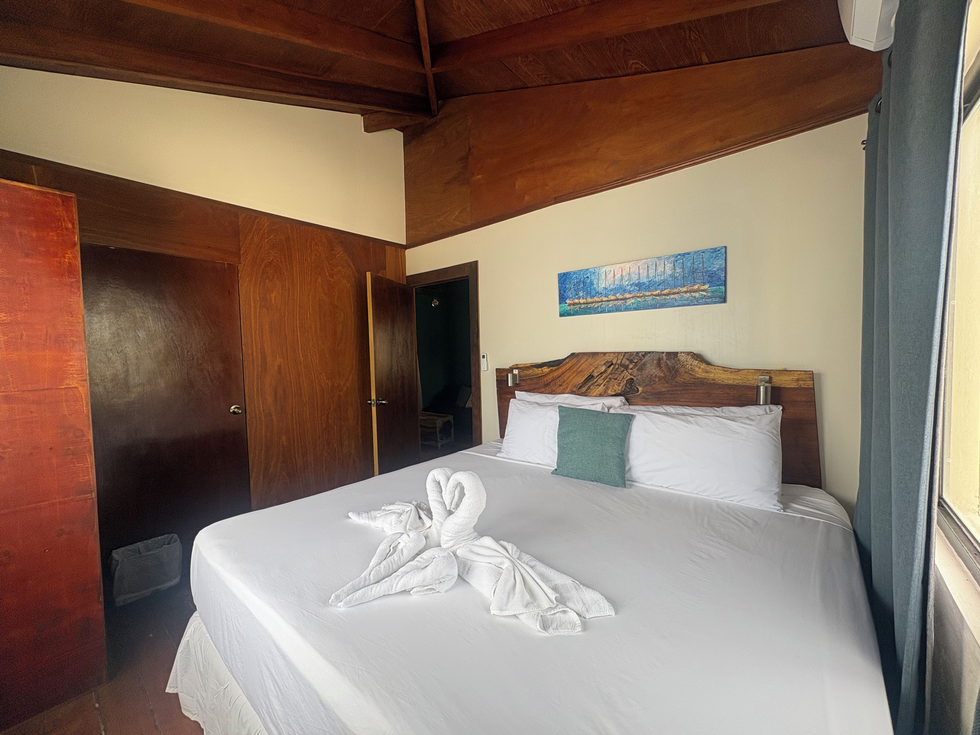 Habitación de hotel con techo de madera, cama con sábanas blancas, un cuadro en la pared y una puerta a la izquierda.