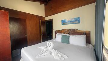 Habitación de hotel con techo de madera, cama con sábanas blancas, un cuadro en la pared y una puerta a la izquierda.