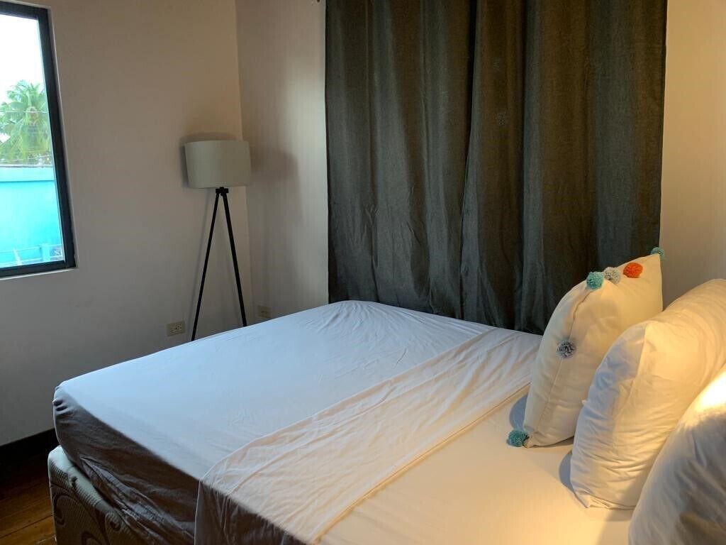 Een net opgemaakt bed met witte lakens, een vloerlamp en uitzicht op een zwembad door een raam.