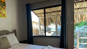 Una cama bien hecha con sábanas blancas y toallas enrolladas, una ventana con vista a una construcción con techo de paja y una cortina azul.
