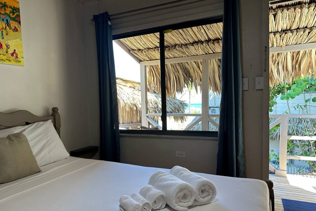 A neatly made bed with white linens and rolled towels, a window with a view of a thatched-roof structure, and a blue curtain.