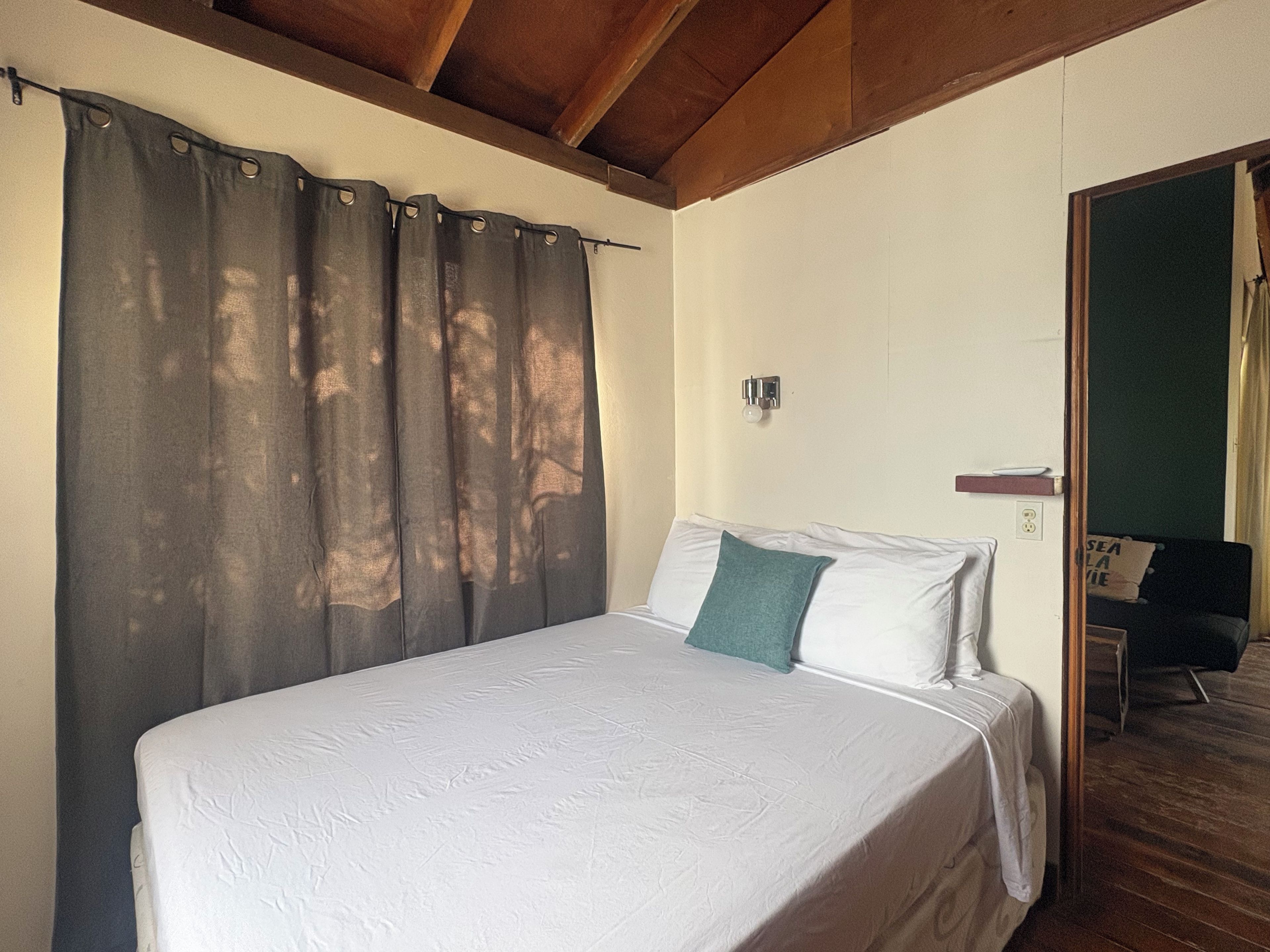 Un lit simple avec un matelas blanc et un oreiller turquoise, un plafond en bois, une lampe fixée au mur et une porte donnant sur une autre pièce.