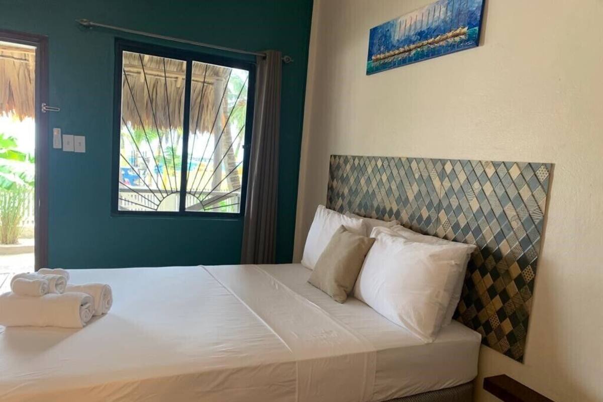A neatly made bed with white linens and pillows, a window with a view of greenery, and a painting on the wall.