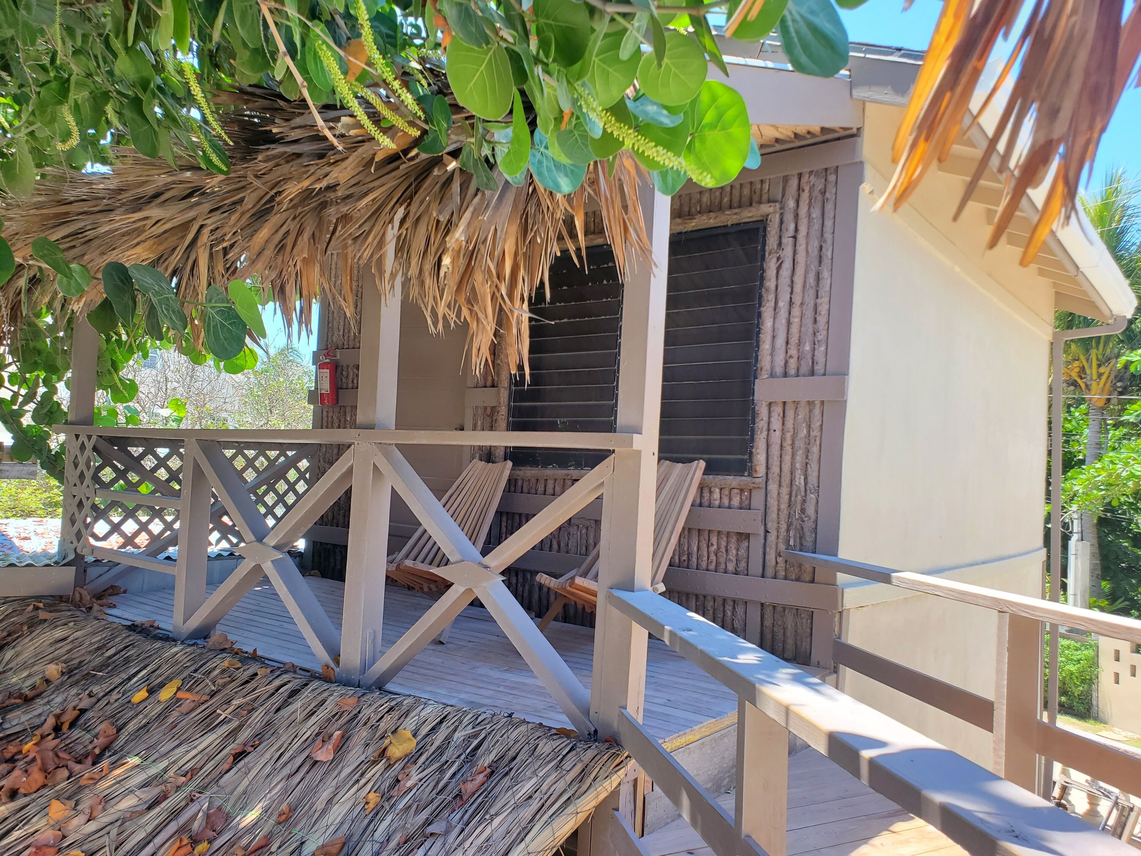 Une terrasse en bois surmontée d’un toit de chaume, avec une balustrade blanche, et d’où l’on peut admirer un bâtiment doté d’une fenêtre et d’une porte.
