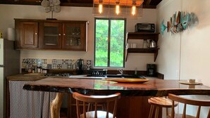 Una cocina con armarios de madera, una isla grande con taburetes, un ventanal con vistas a follaje, y un estante en la pared con objetos decorativos.