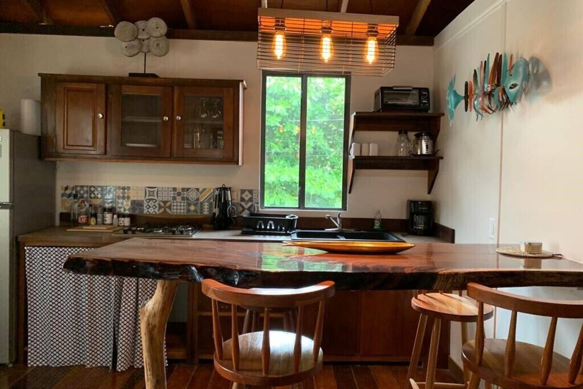 A kitchen with wooden cabinets, a large island with stools, a window with a view of greenery, and a wall-mounted shelf with decorative items.