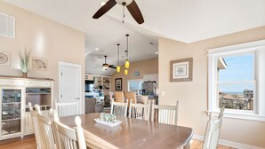A dining area with a wooden table and chairs, a ceiling fan, pendant lights, and a view of the city through a large window. - Another Day In Paradise: 4-Min Walk to the Beach! (Kill Devil Hills)
