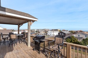 Terraza de madera con mobiliario exterior y una parrilla, con vistas a una zona residencial y al océano.