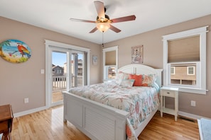 A bedroom with a bed, ceiling fan, wooden floor, and a view of the outside.