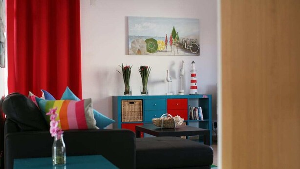 A living room with a sofa, a teal coffee table, and a red curtain. There is a painting on the wall and a white lamp on a blue cabinet. - Villa Ocenano  - Stunning Ocean Views (Albufeira)