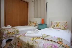 Une chambre d’hôtel avec deux lits simples, chacun agrémenté d’une literie à motifs floraux et d’une tête de lit blanche. On y trouve une armoire en bois et une petite table de chevet de couleur turquoise avec une lampe.