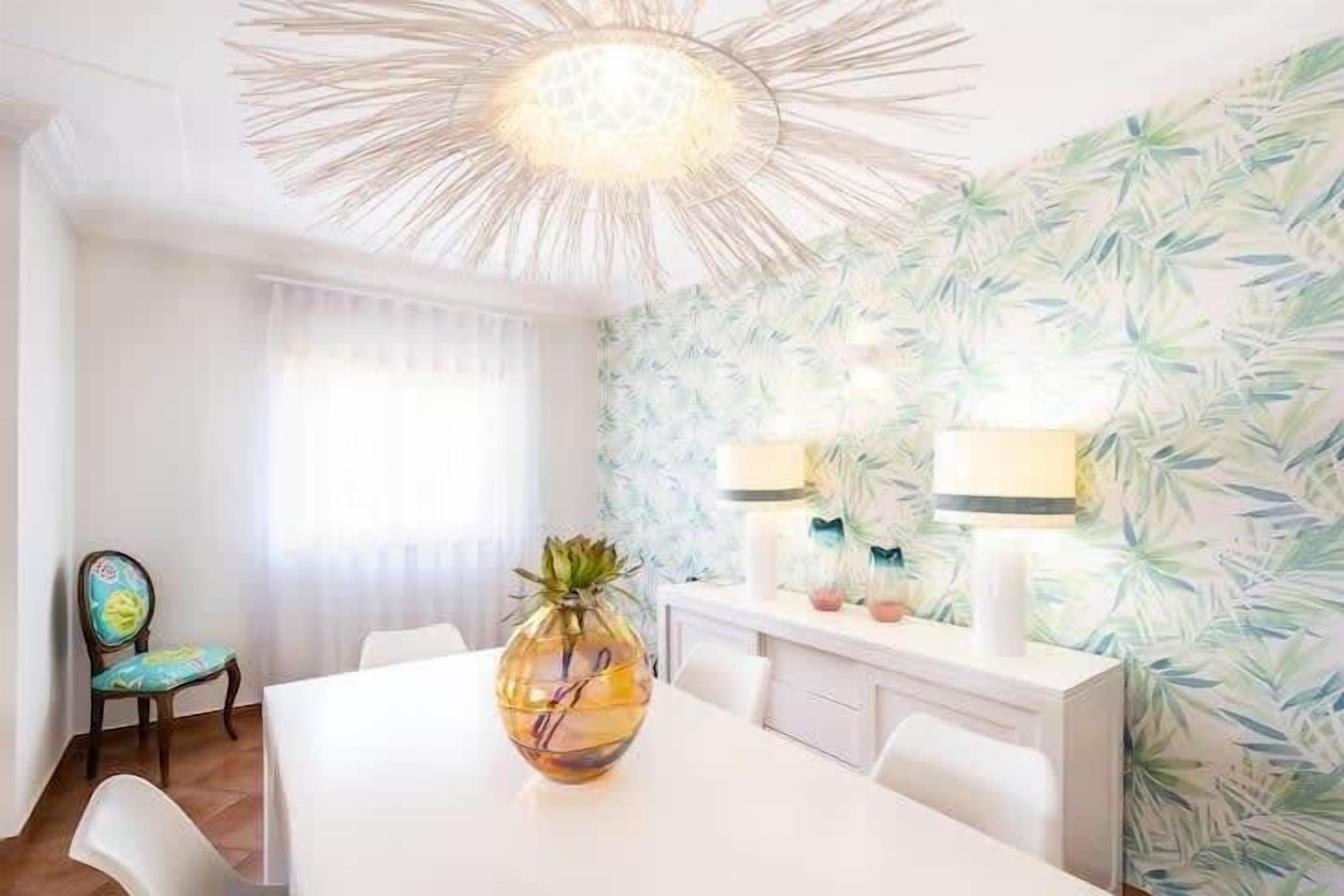Un comedor con una mesa blanca, sillas, un jarrón decorativo y una pared con diseño de hojas.