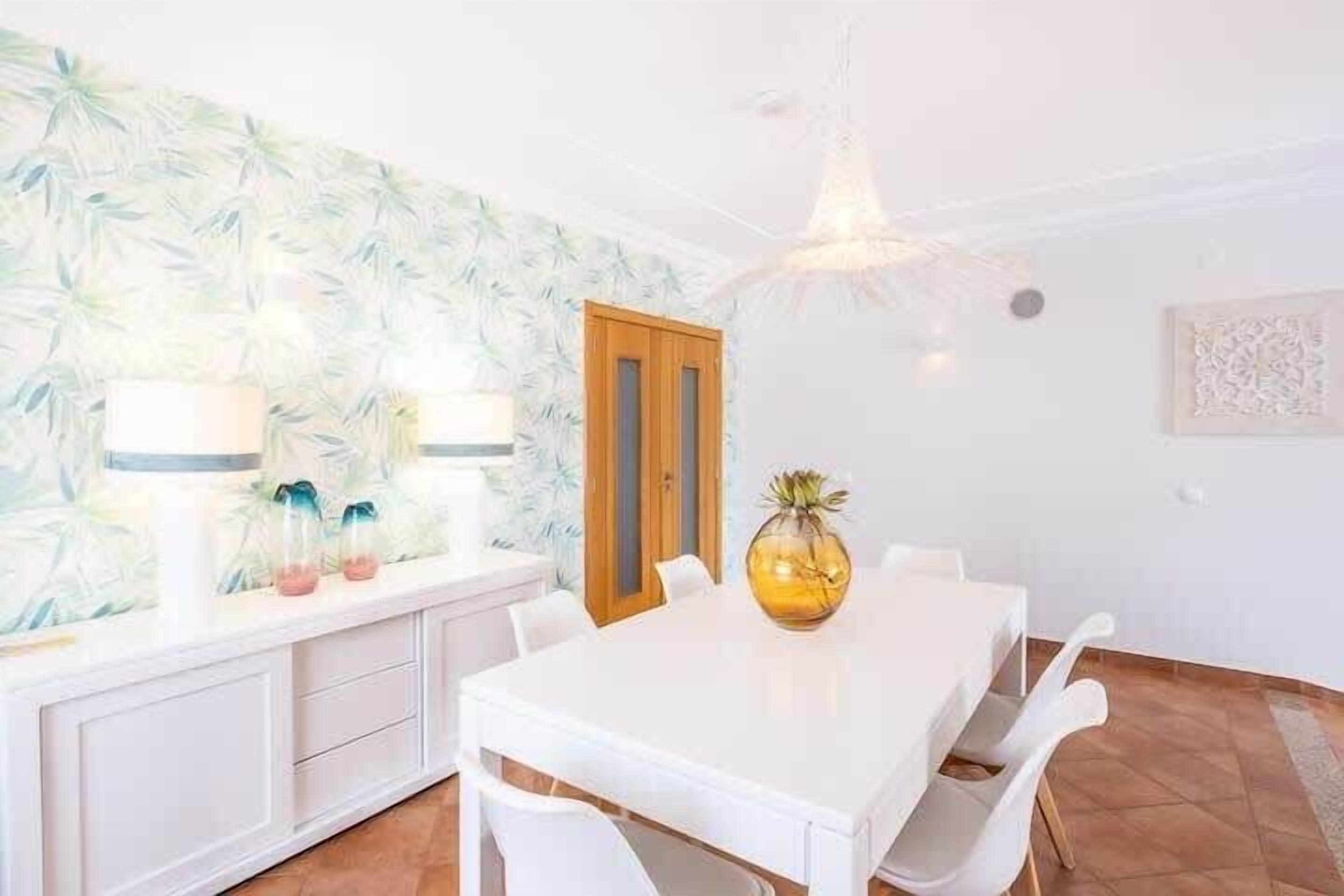 Un coin repas avec une table et des chaises blanches, un vase avec une plante et un mur orné d’un motif végétal.