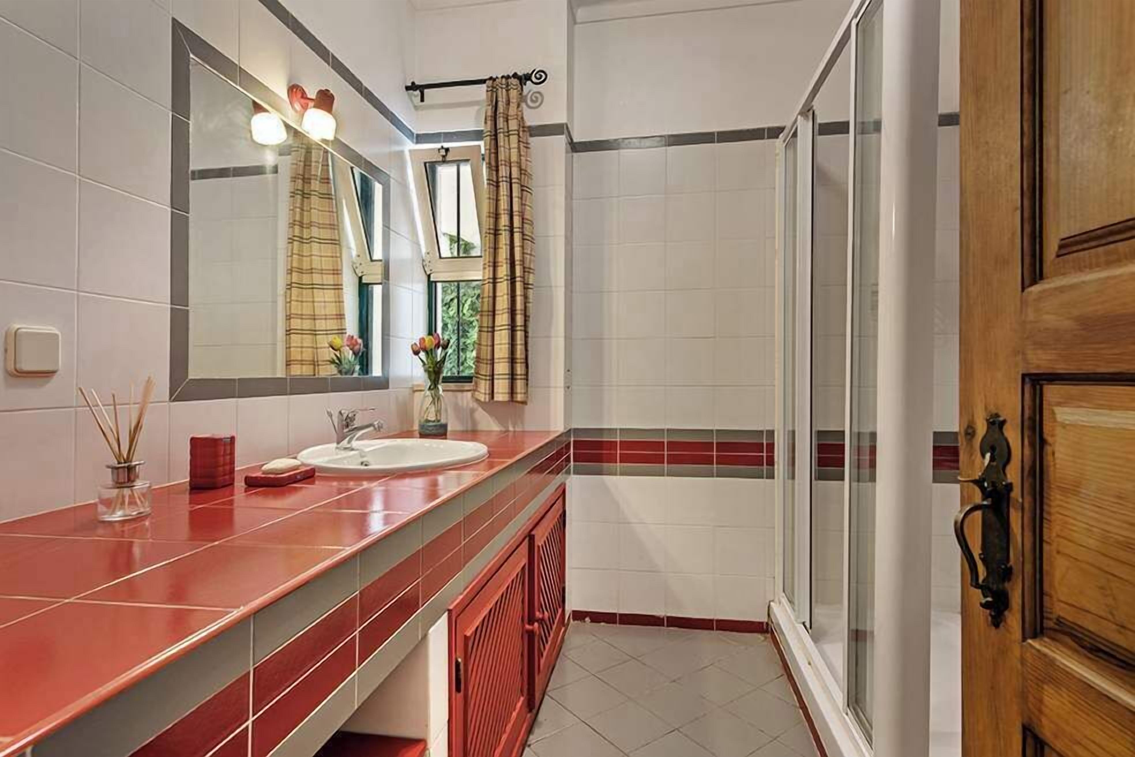 Un baño con encimera roja, un espejo, un lavabo, ducha y ventana con vistas.