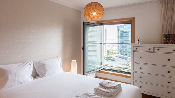 Une chambre d’hôtel avec un lit, une commode, une fenêtre donnant sur des immeubles et un luminaire suspendu.