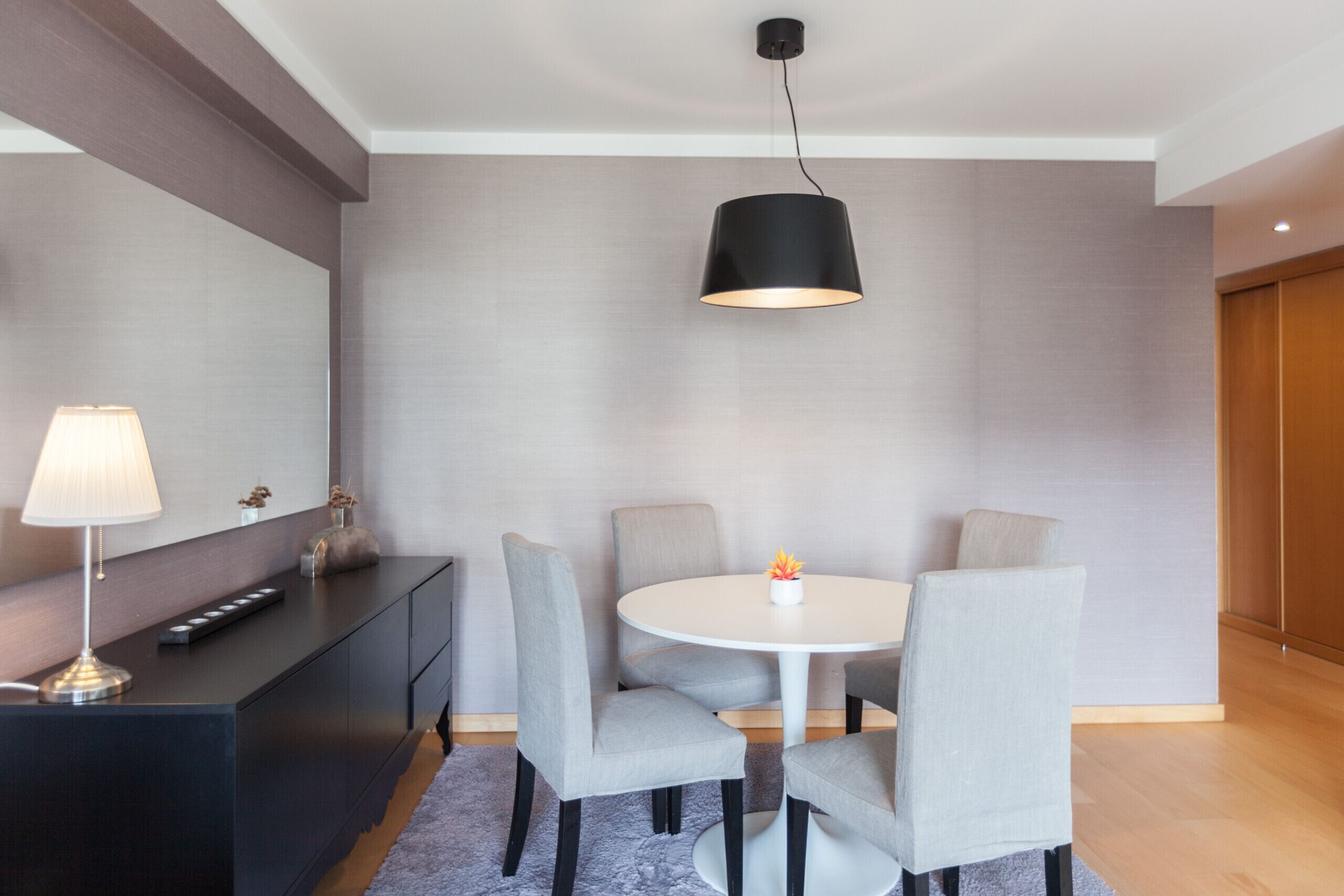 A modern dining area with a round table, grey upholstered chairs, a black pendant light, and a dark wooden console with a lamp.