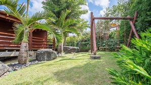 A tropical garden with a wooden swing set, palm trees, and a rustic building.