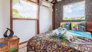 A bedroom with a bed, a wooden nightstand, a stone wall, and a window with a view of greenery.