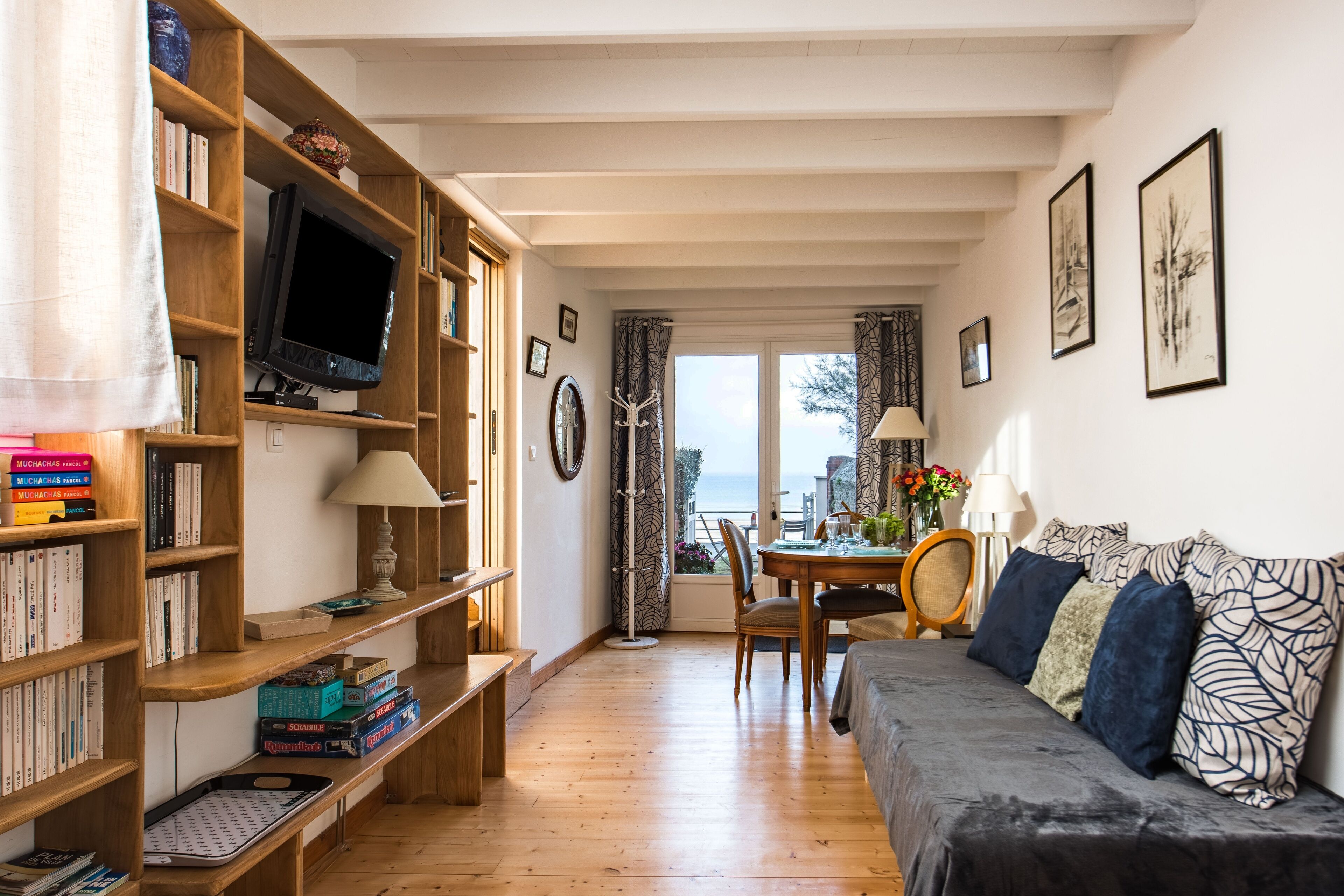 A cozy living room with a wooden bookshelf, a flat-screen TV, a sofa with patterned cushions, and a dining area with a table and chairs.