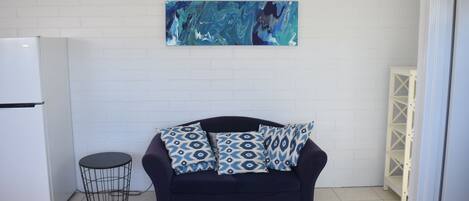Une pièce avec un mur en briques blanches, un canapé bleu foncé orné de coussins à motifs, une petite table ronde et un réfrigérateur blanc.