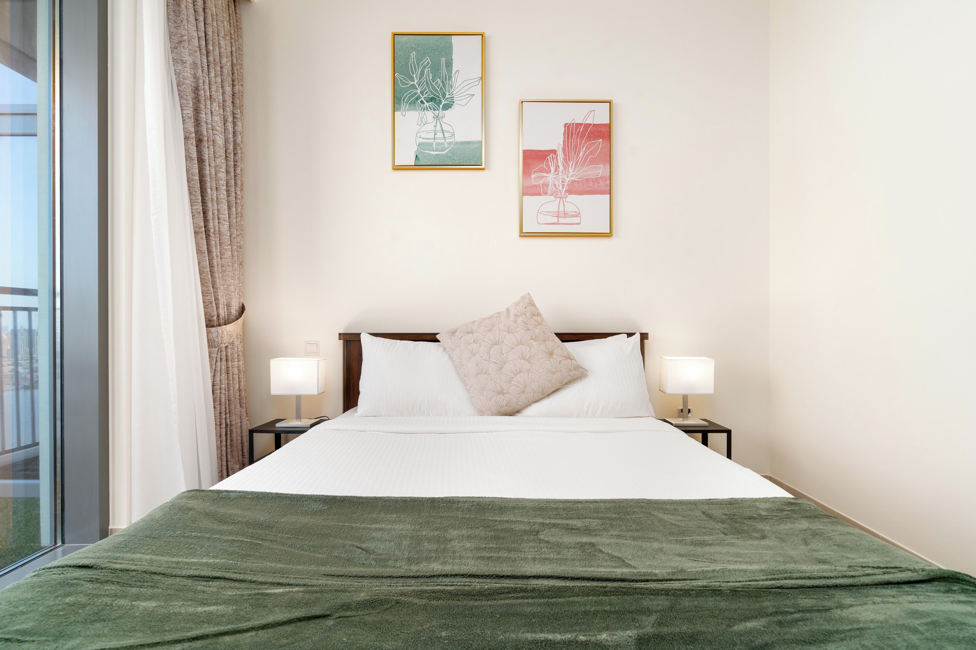A neatly made bed with a green blanket, two bedside lamps, and a pillow on a white mattress. There are two framed artworks on the wall above the bed.